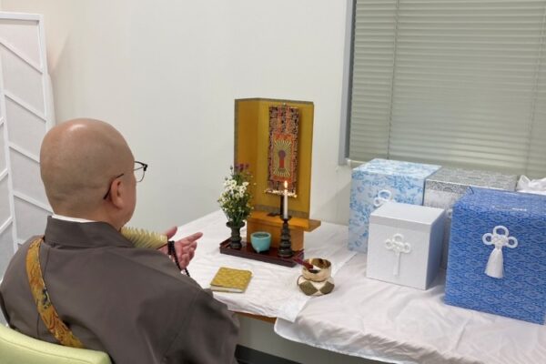 ライフジェムの事務所の一角で僧侶が読経し、骨壺の供養を行っている様子。供養台には骨壺が置かれている。